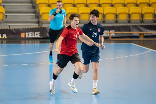 Jakub Smoliński w czerwonej koszulce z numerem 14 biegnie z piłką ręczną, obok zawodnik w granatowej koszulce z numerem 29.