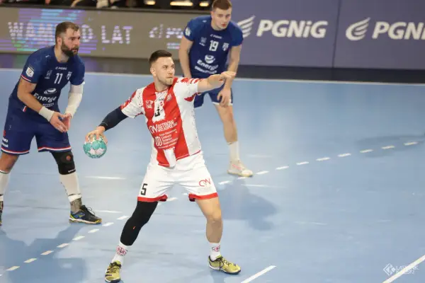 Kamil Adamski w akcji, rzuca piłkę do bramki. Zawodnik w białej koszulce z czerwonymi pasami i numerem 5.