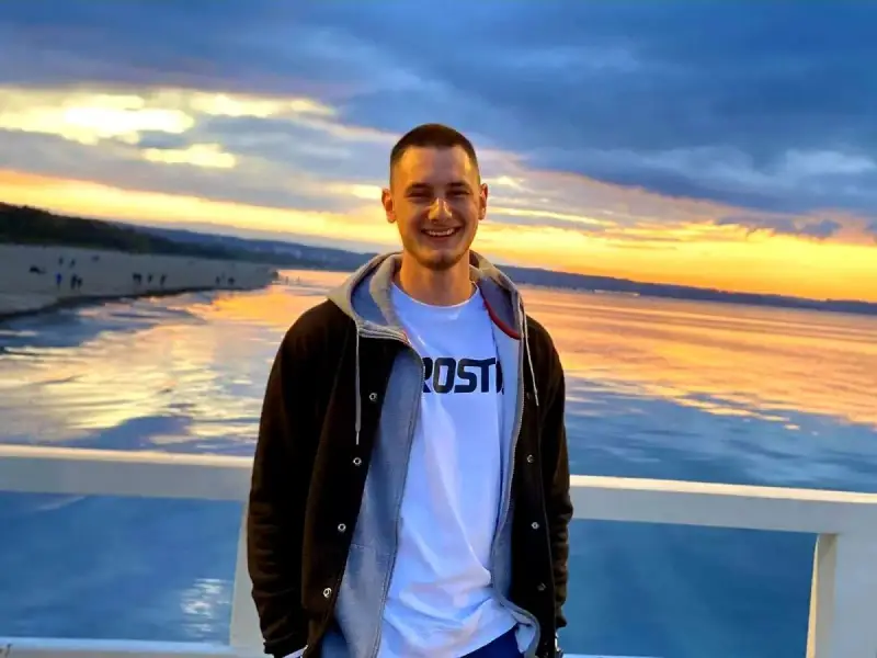 Uśmiechnięty Krzysztof Komarzewski na molo o zachodzie słońca. W tle plaża i morze.