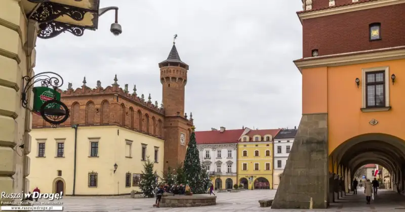 Rynek w Tarnowie zimą, z choinką i ludźmi przy fontannie. W tle zabytkowe kamienice i wieża ratuszowa.