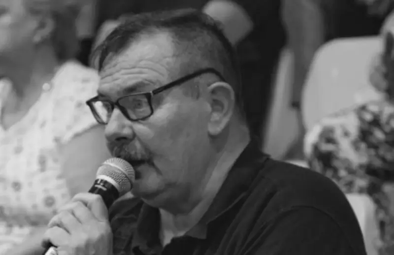 Krzysztof Janicki w okularach i z wąsami mówi do mikrofonu, w tle widać ludzi.